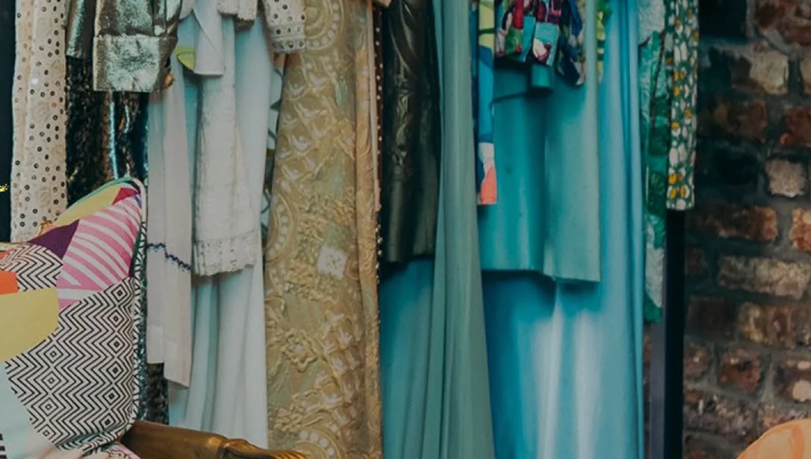 Inside the shop — patterned pillows on a bench, brick wall, racks of dresses